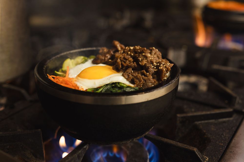 Korean bibimbap in a hot stone bowl with egg, beef, and vegetables cooking over an open flame.