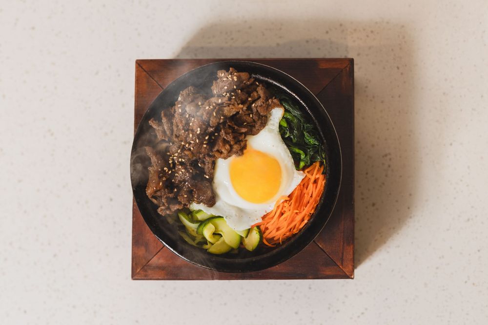 Steaming Korean bibimbap bowl with fried egg, beef, carrots, spinach, and cucumber on a stone hotpot.