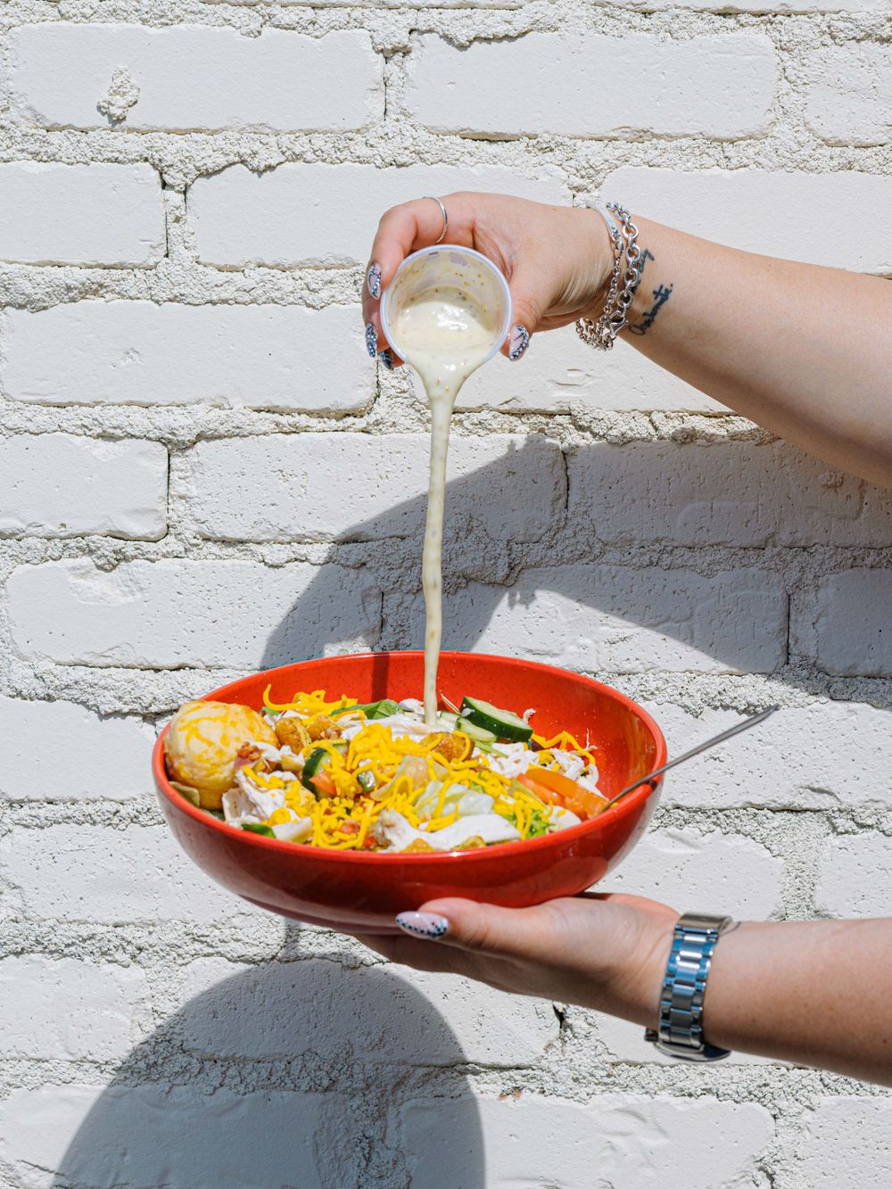 Hands pouring creamy dressing over a vibrant red bowl filled with rice, vegetables, and toppings against a white brick wall.