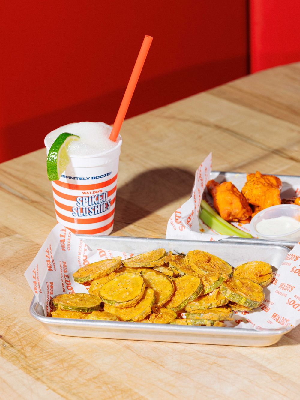 Fried pickle chips, buffalo wings with ranch, and a spiked slushie with lime on a wooden table.