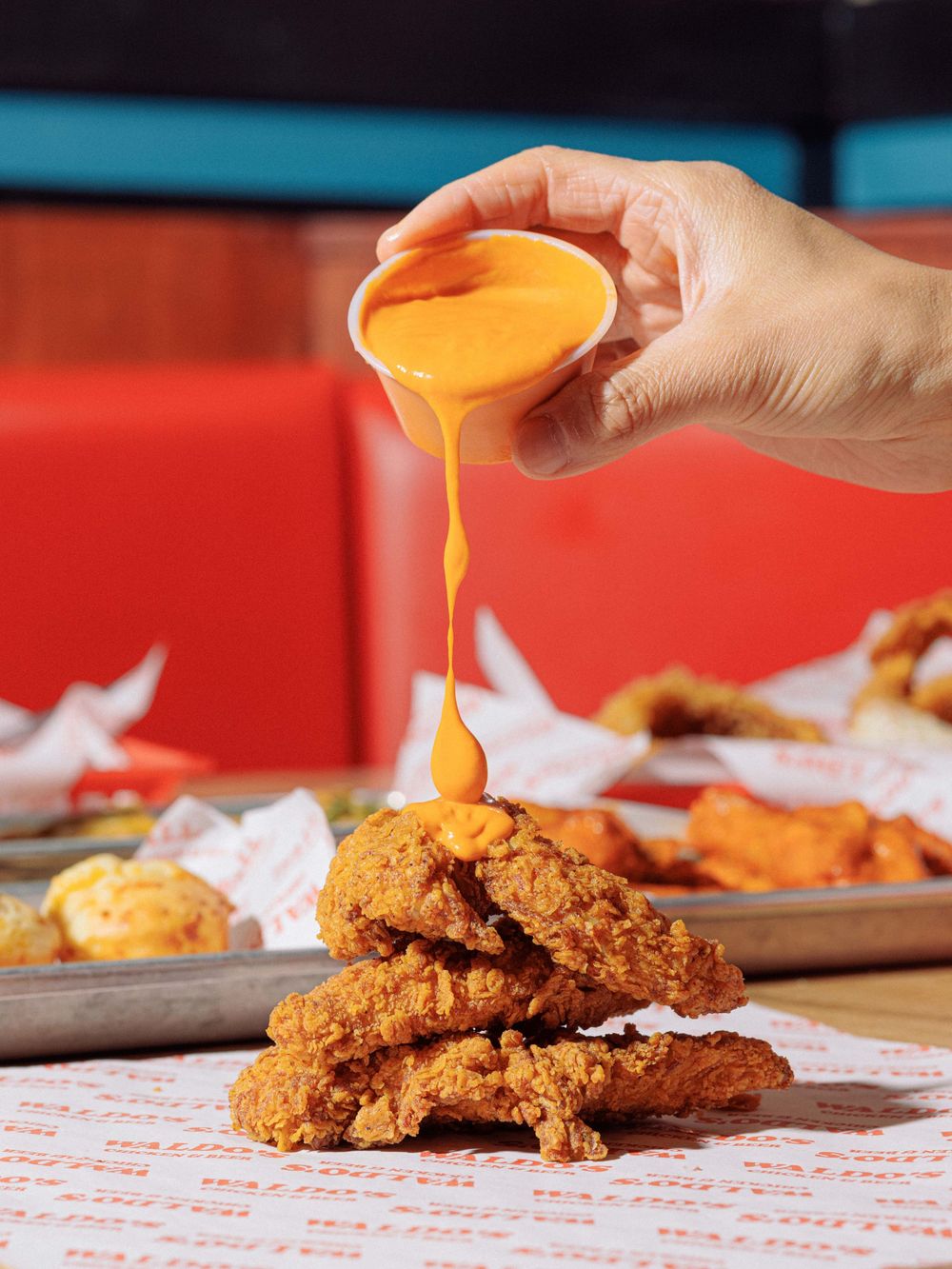 Hand pouring bright orange cheese sauce over crispy fried chicken tenders stacked on a checkered paper.