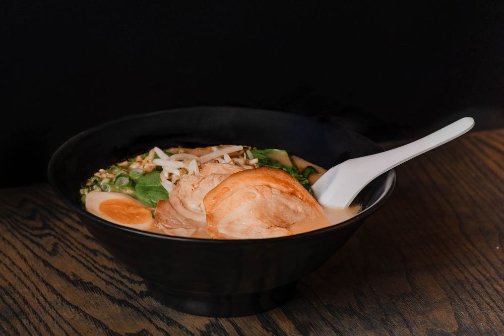 A steaming bowl of Japanese ramen with chashu pork, soft-boiled egg, greens, and noodles in rich broth, served with a white spoon.