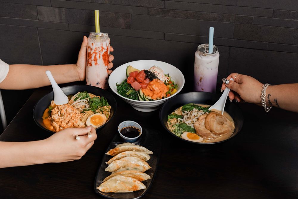 Hands reaching for Asian ramen bowls, dumplings, and bubble tea drinks on a dark restaurant table.