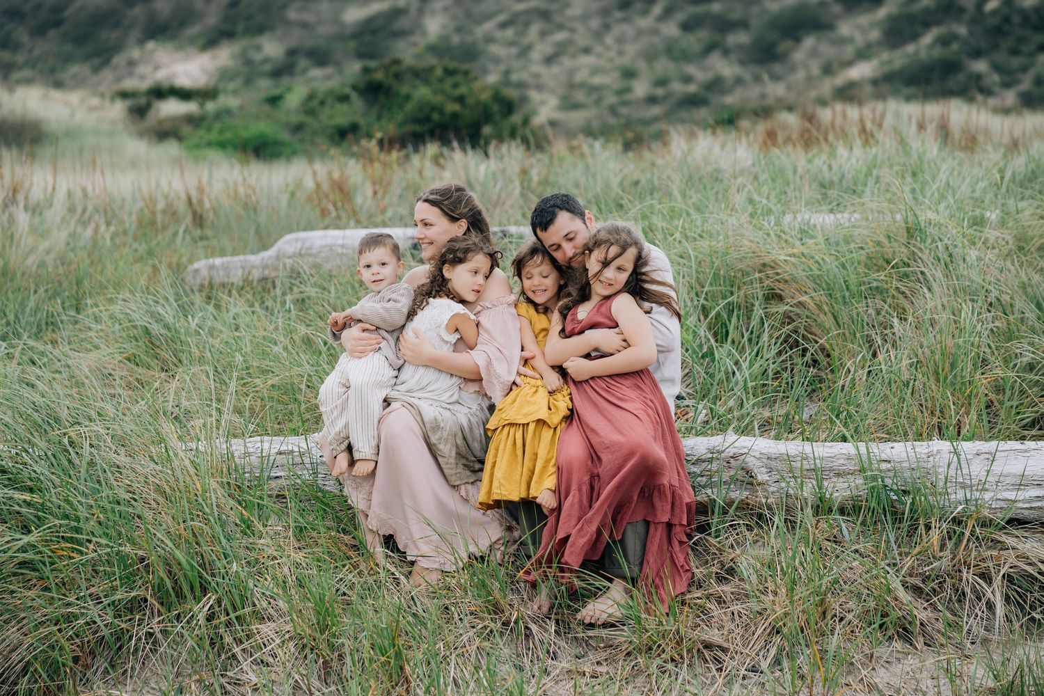 Family of six cuddles together on driftwood log in grassy coastal dunes, children in colorful dresses embracing parents.