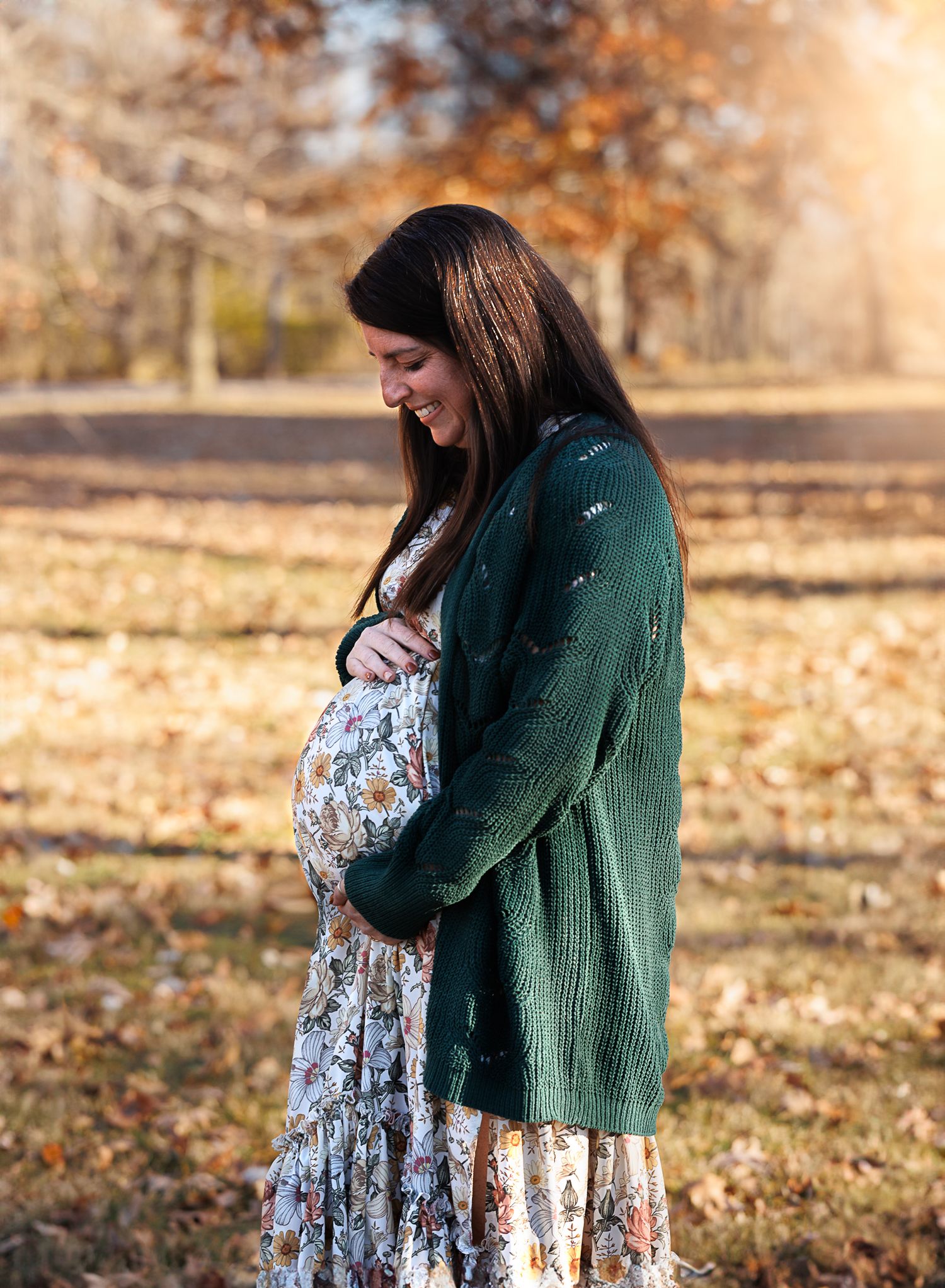 Smiling pregnant woman in floral dress and green cardigan cradling her baby bump outdoors in autumn park.