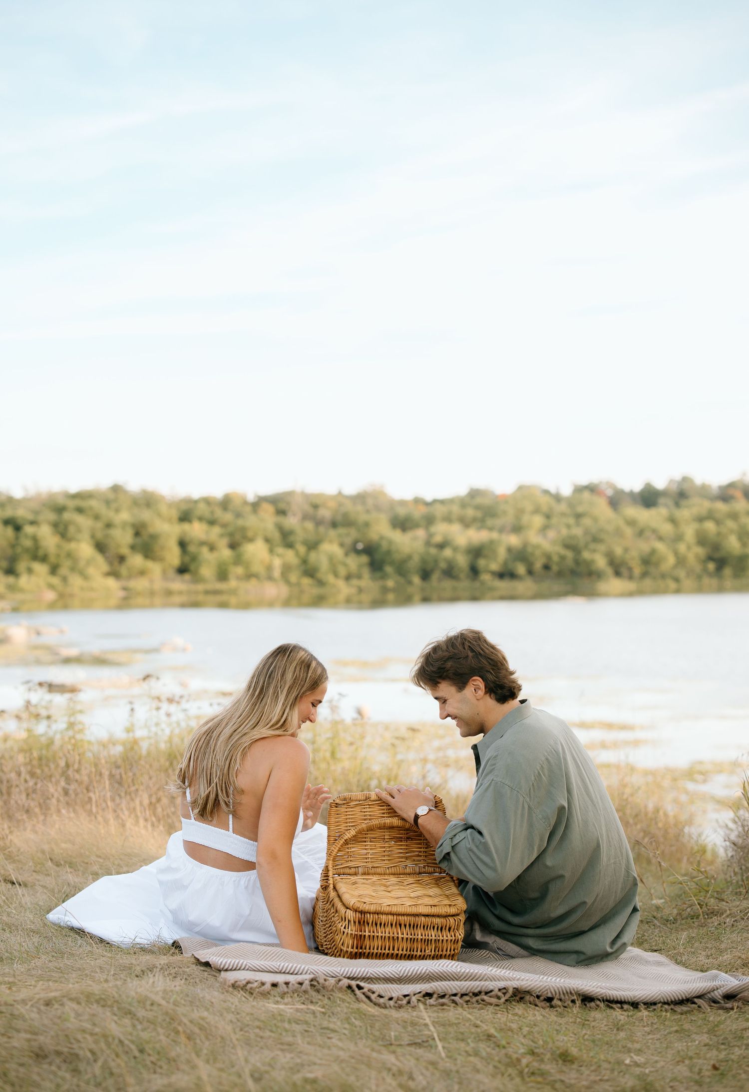 Couple sharing a romantic picnic by a serene lake, sitting on a blanket with a wicker basket in a golden field.