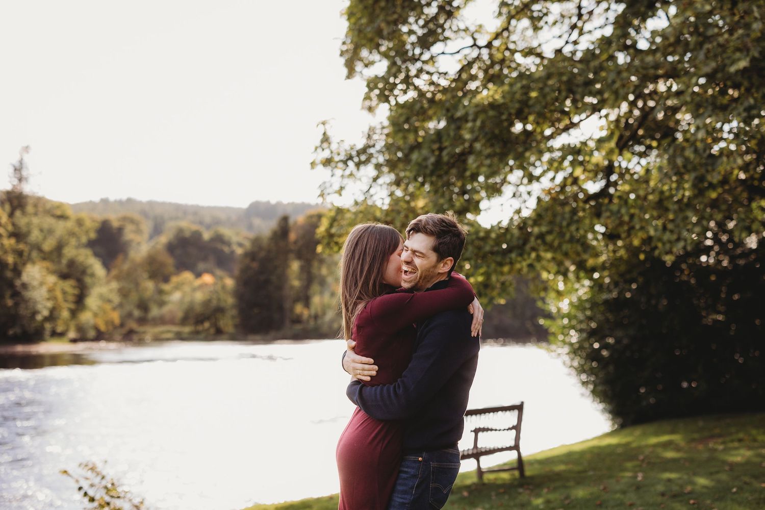 Couple embracing and laughing by a scenic riverside park with lush green trees on a sunny day.