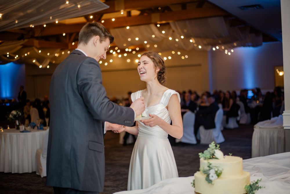 Bride and groom laughing together during wedding cake cutting ceremony in elegant reception hall with string lights.