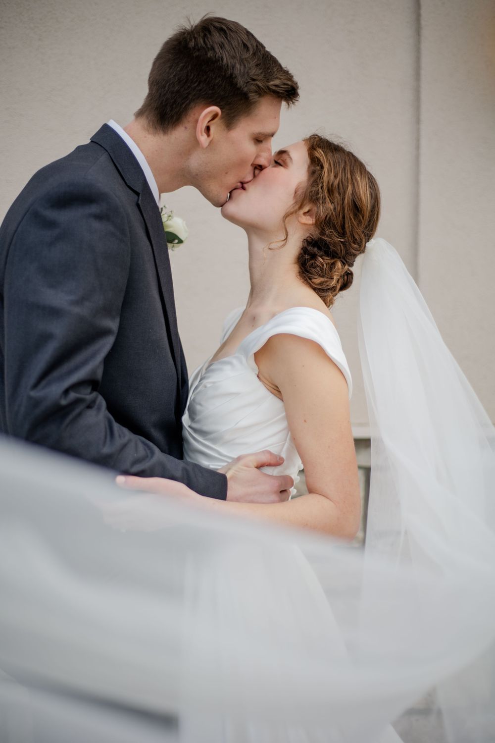 Bride and groom sharing a romantic first kiss, with wedding veil flowing in the wind during outdoor ceremony.