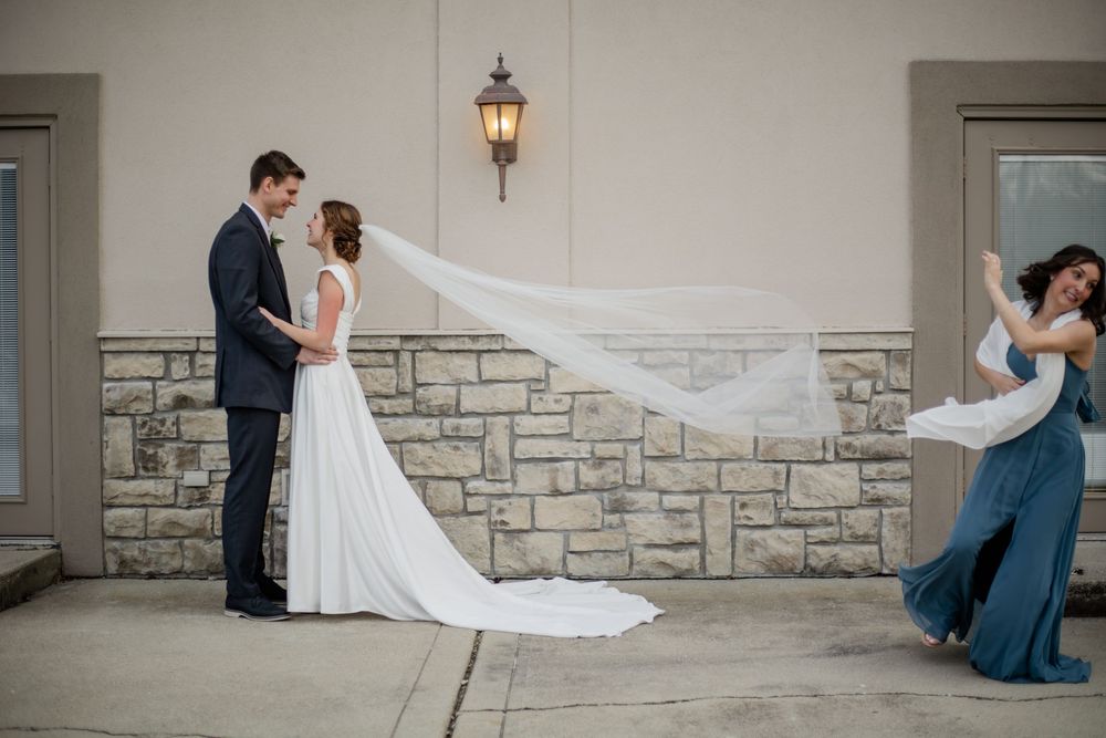 Bride and groom share a romantic moment outdoors while a bridesmaid in blue holds the flowing wedding veil.