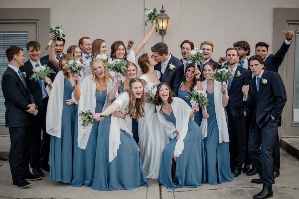 Wedding party in blue dresses and dark suits celebrating outdoors, bride and groom kissing in center.