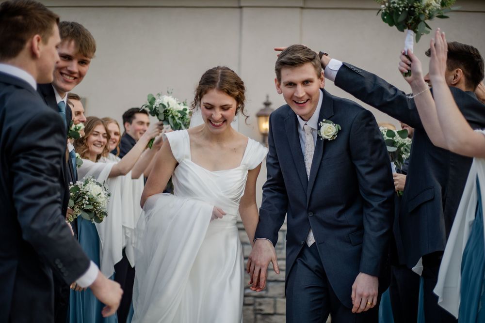 Joyful bride and groom walk through a celebratory arch of raised bouquets surrounded by smiling wedding party guests.