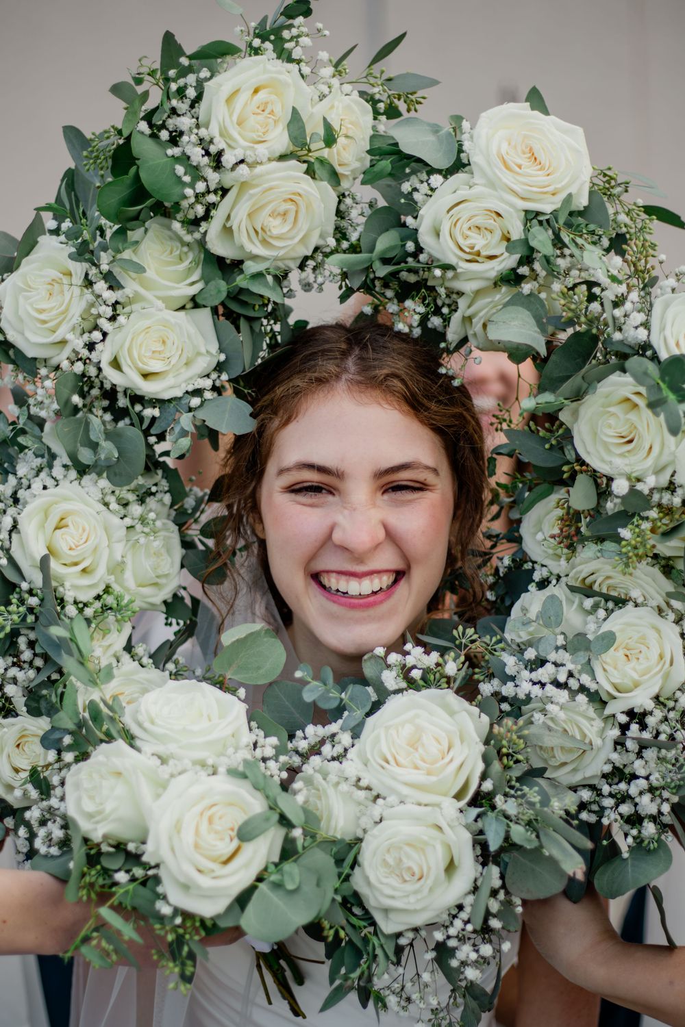 Joyful bride laughing through a circle of white rose and eucalyptus bouquets on her wedding day.