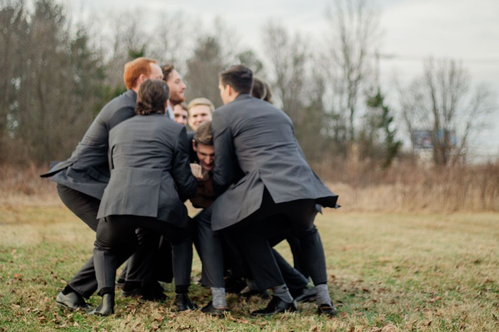 Groomsmen in dark suits playfully huddle together in a grassy field during an outdoor wedding celebration.