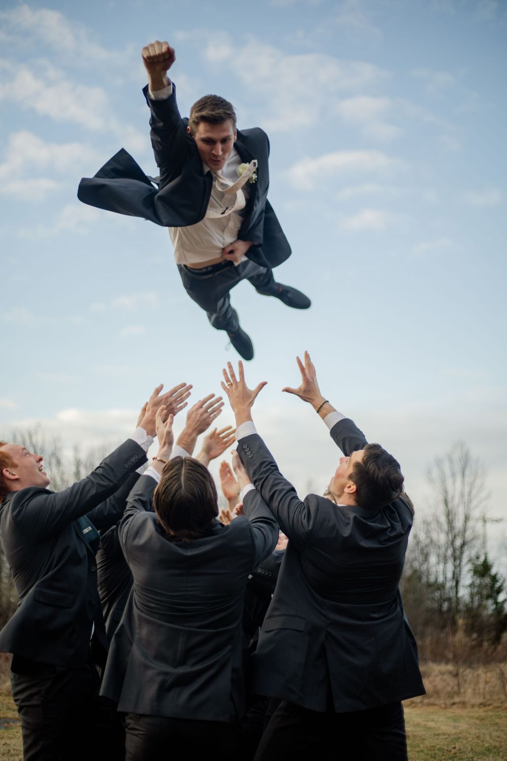Groomsmen tossing groom in the air outdoors during wedding celebration under a blue sky.