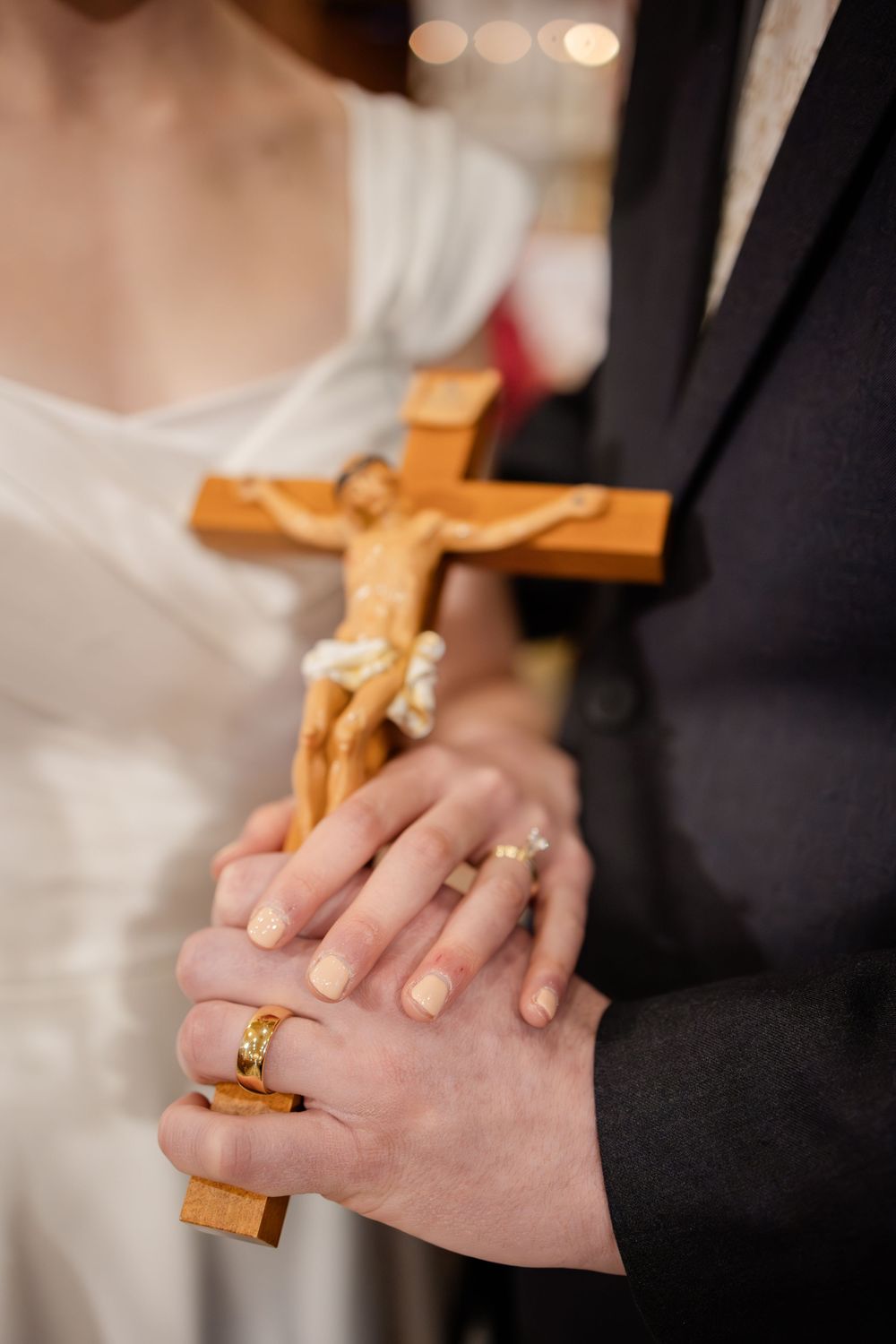 Bride and groom holding a wooden crucifix together, showing their gold wedding rings during a religious ceremony.