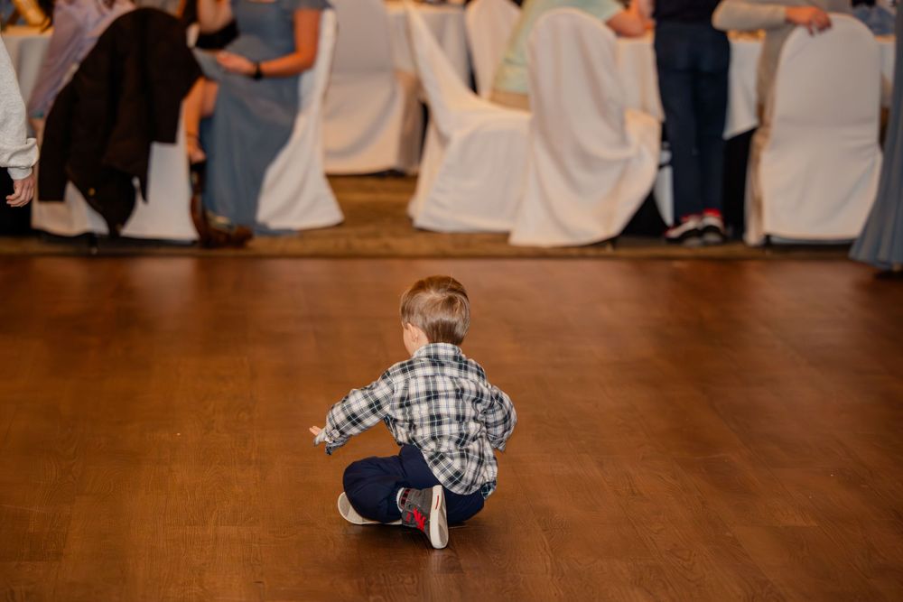 Young child sitting on wooden dance floor at wedding reception with guests seated in background.