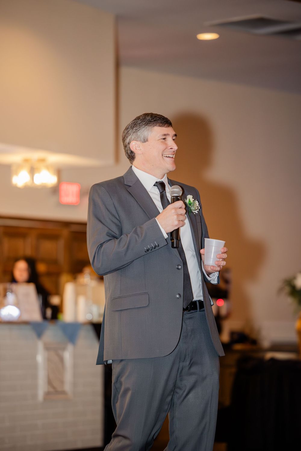 Man in gray suit giving a toast with microphone at a wedding reception.