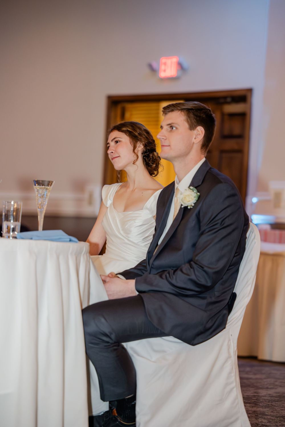 Bride and groom sitting together at sweetheart table during wedding reception, dressed in white gown and navy suit.
