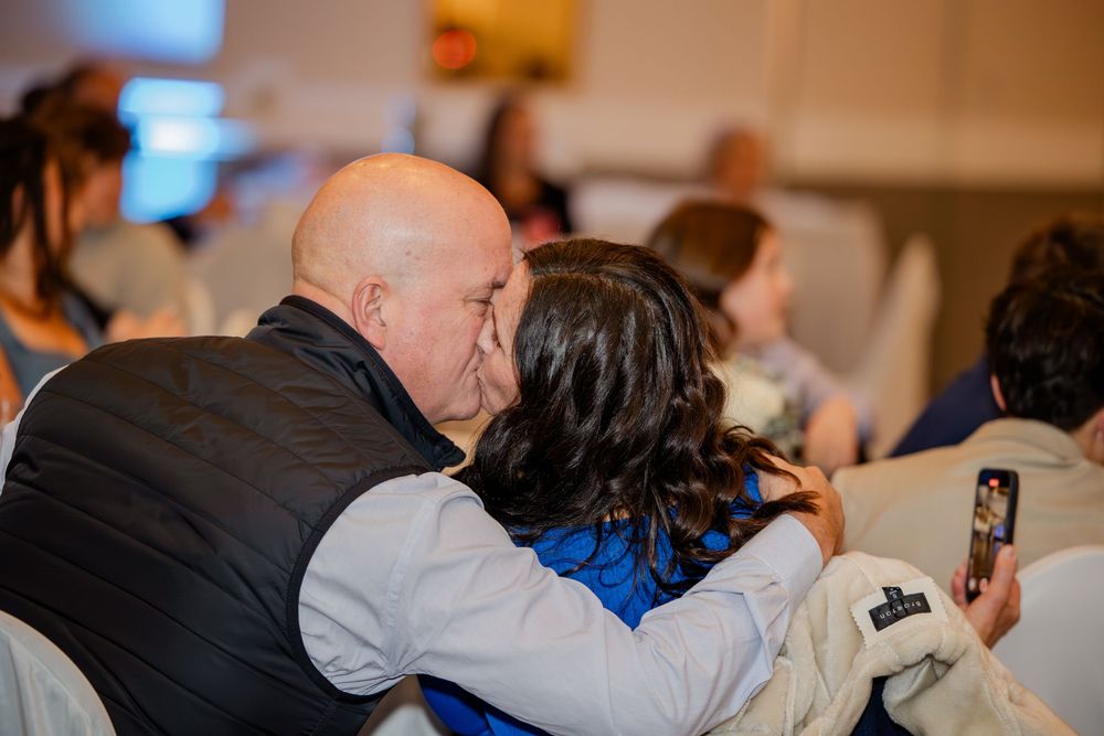 A bald man and a woman sharing a kiss at an indoor event, with guests visible in the background.
