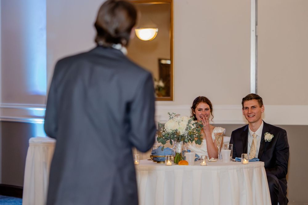 A smiling bride and groom sit at the sweetheart table while a guest delivers a wedding speech.
