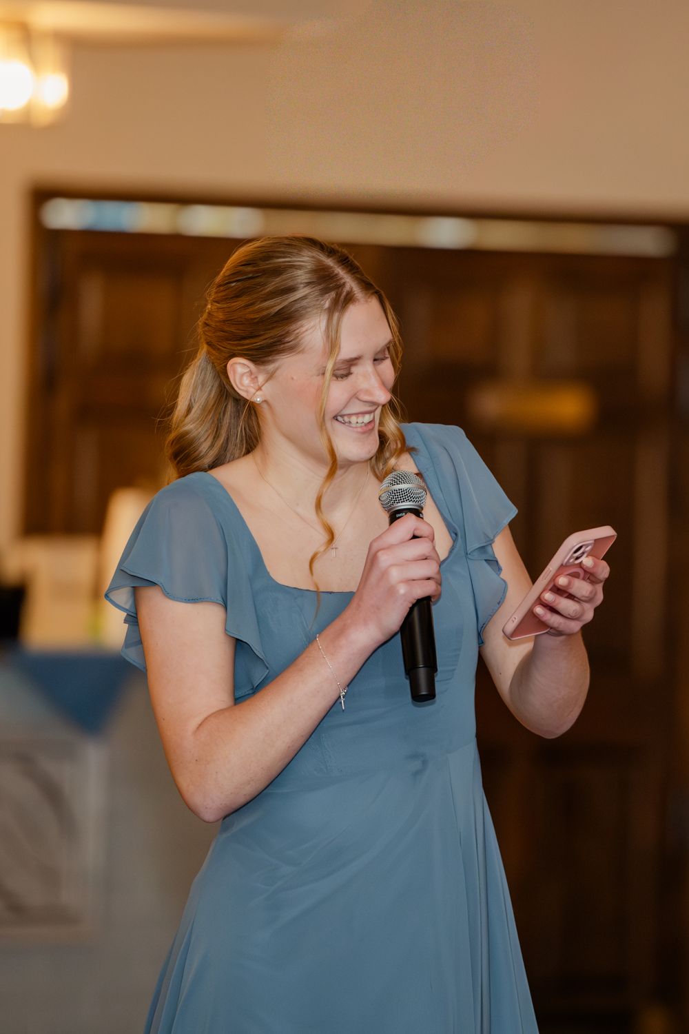 Bridesmaid in blue dress giving a speech, holding microphone and phone, smiling at a wedding reception.