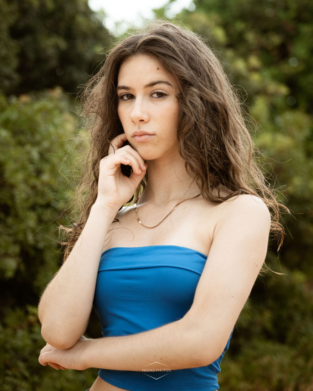 Young woman in blue tube top posing outdoors with hand on chin, surrounded by lush green foliage.