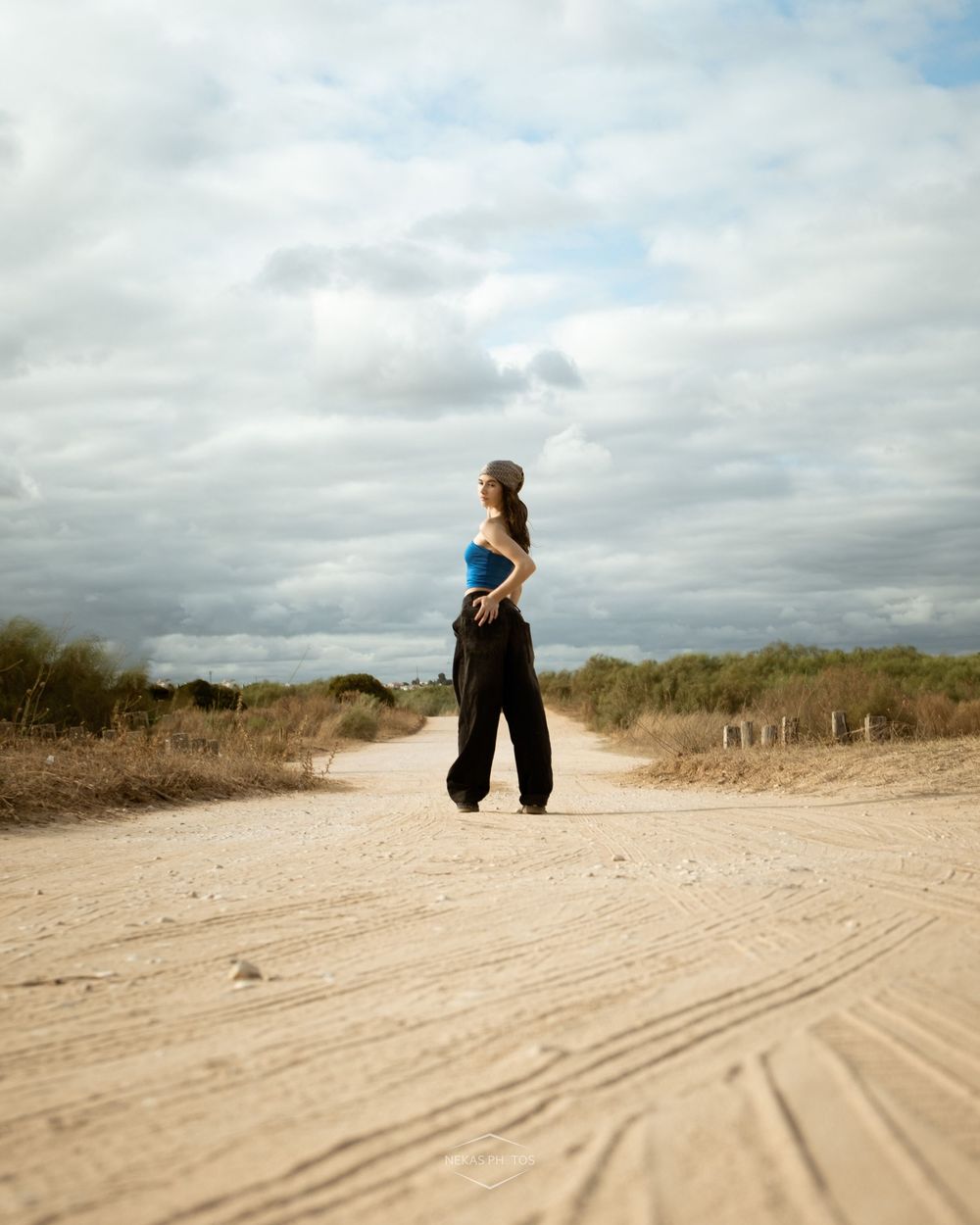 Woman in blue top and black pants standing on a sandy dirt road under a dramatic cloudy sky.