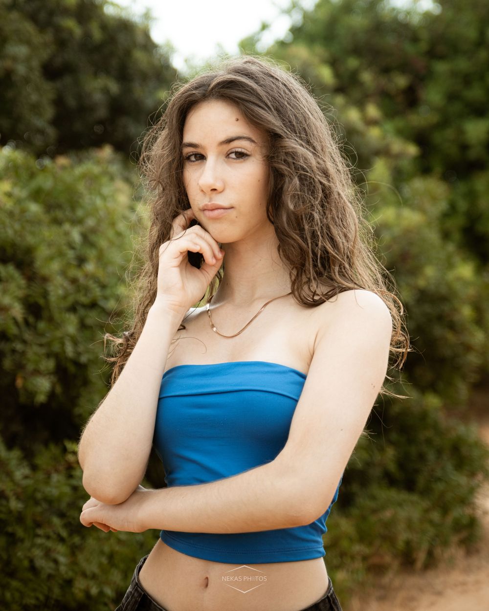 Young woman in blue tube top poses thoughtfully with hand to chin against a green leafy background.