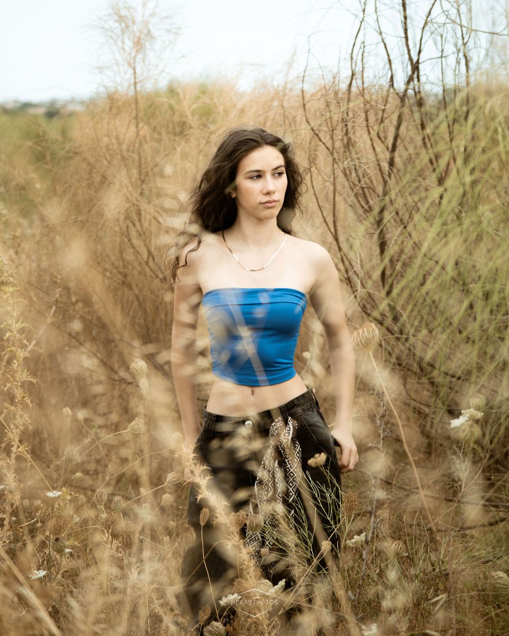 Young woman in blue tube top standing among tall dry grass in an outdoor field setting.