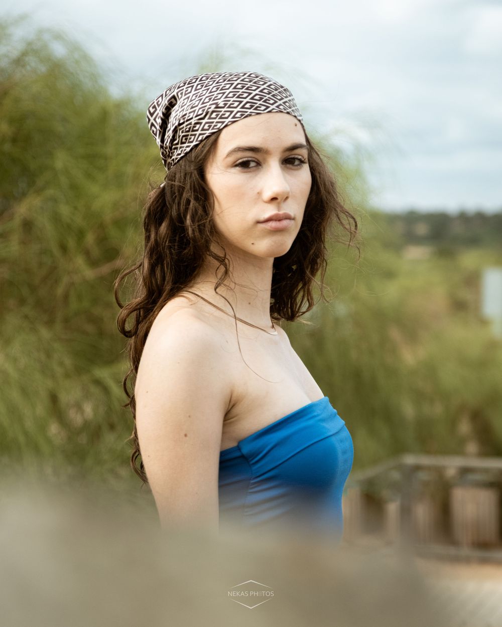 Young woman in blue strapless top and patterned headscarf poses outdoors among green foliage.