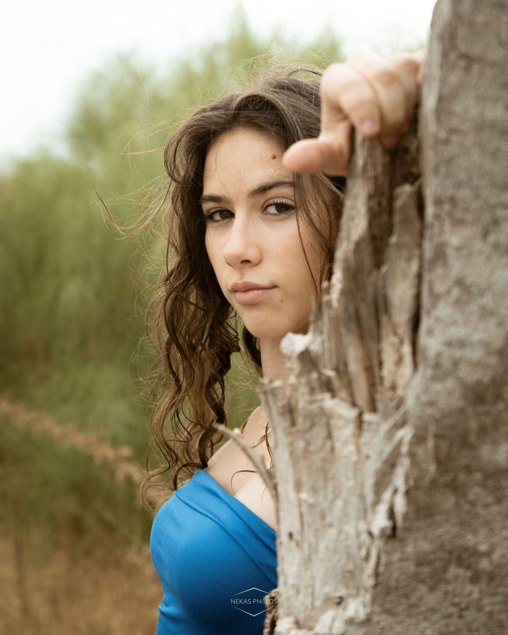 Young woman with curly brown hair in a blue top peers around a weathered tree trunk outdoors.