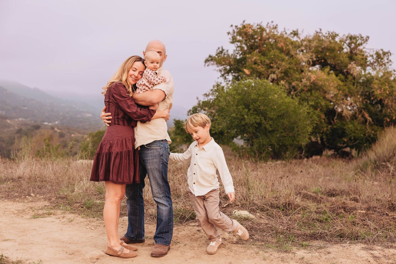 Family of four posing outdoors on a dirt path with green trees and misty hills in the background.