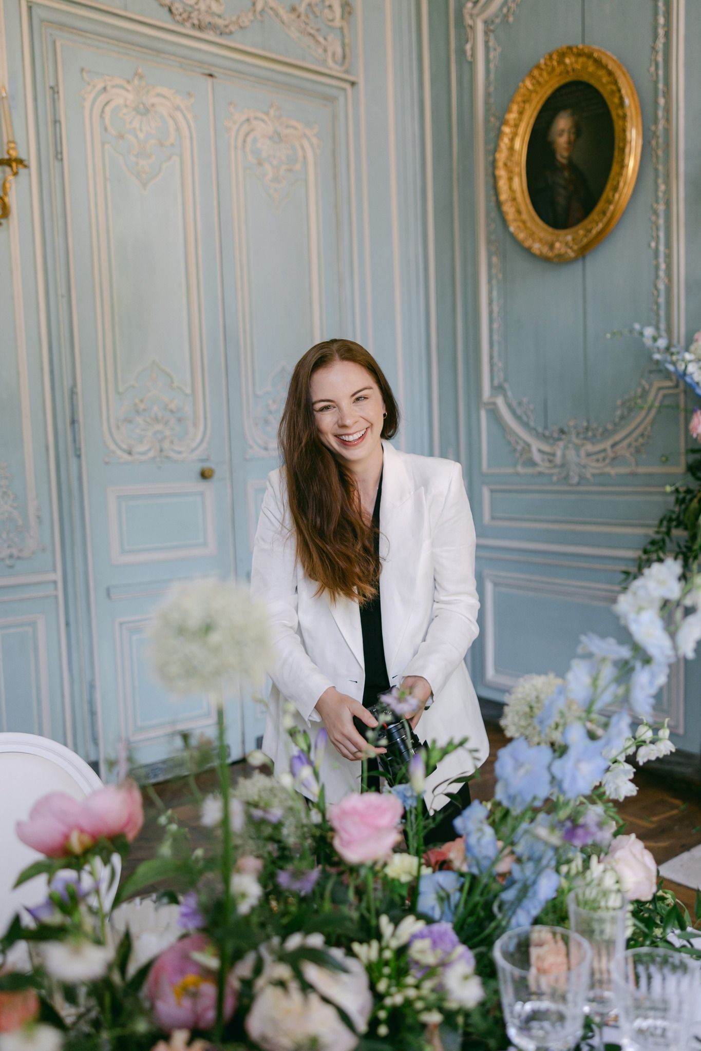 Smiling woman in white blazer arranging colorful flowers in an elegant blue French-style room with gold portrait.