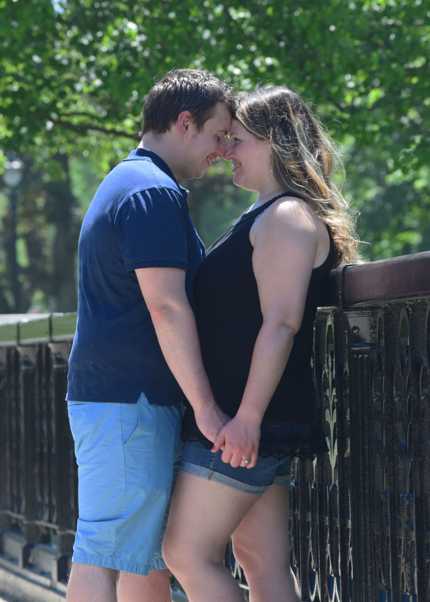 A couple shares a romantic moment on a bridge, holding hands and leaning in for a kiss outdoors.