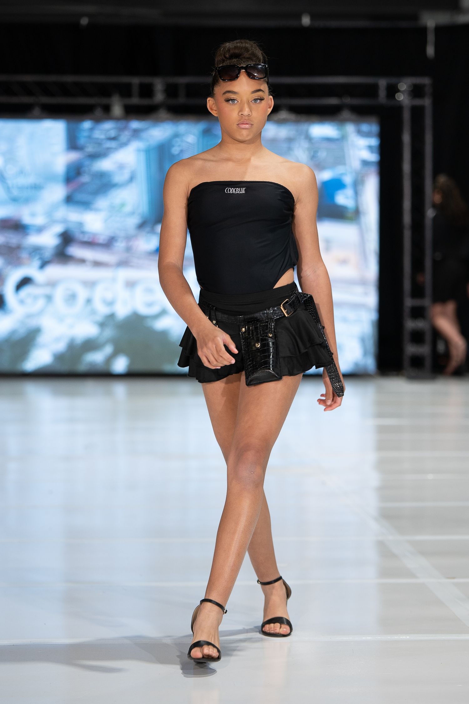 Model walks runway in black strapless top, mini skirt, and heeled sandals at a fashion show.