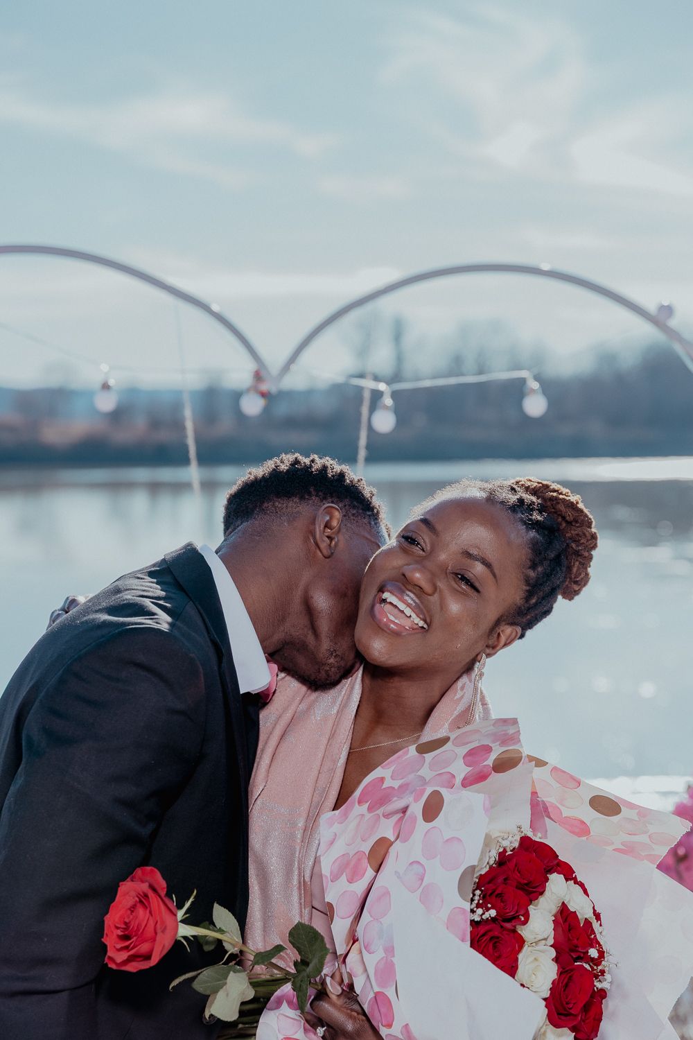 A joyful couple shares a romantic moment outdoors, man kissing woman's cheek as she laughs holding red roses.