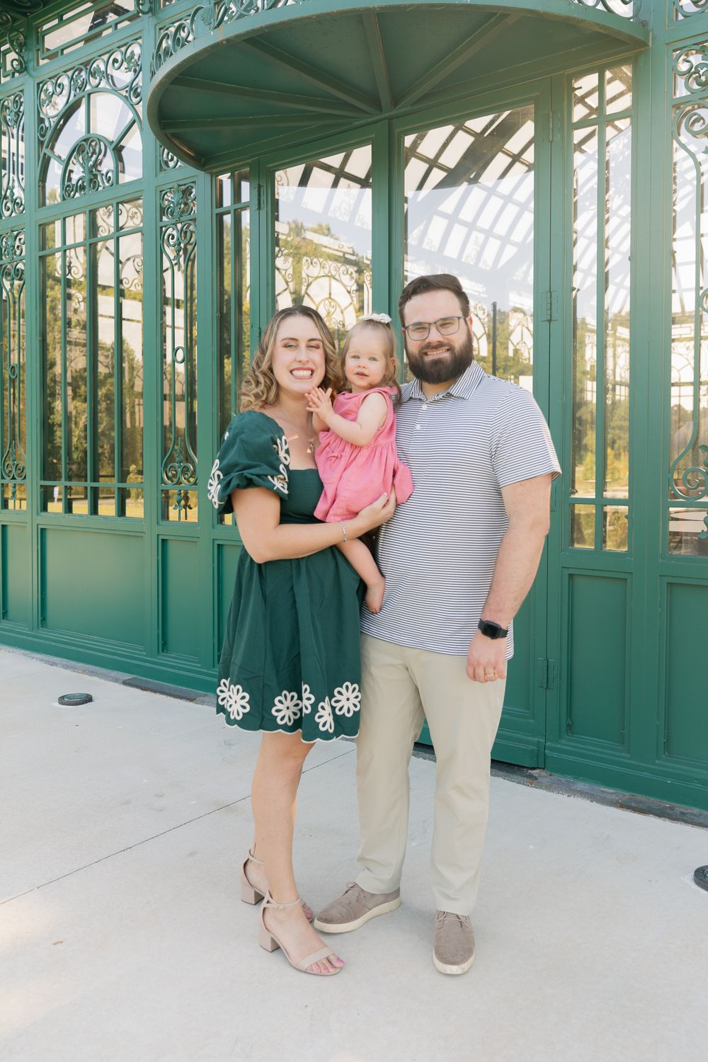 A smiling family of three poses before a green ornate gazebo, with parents holding their toddler in a pink dress.