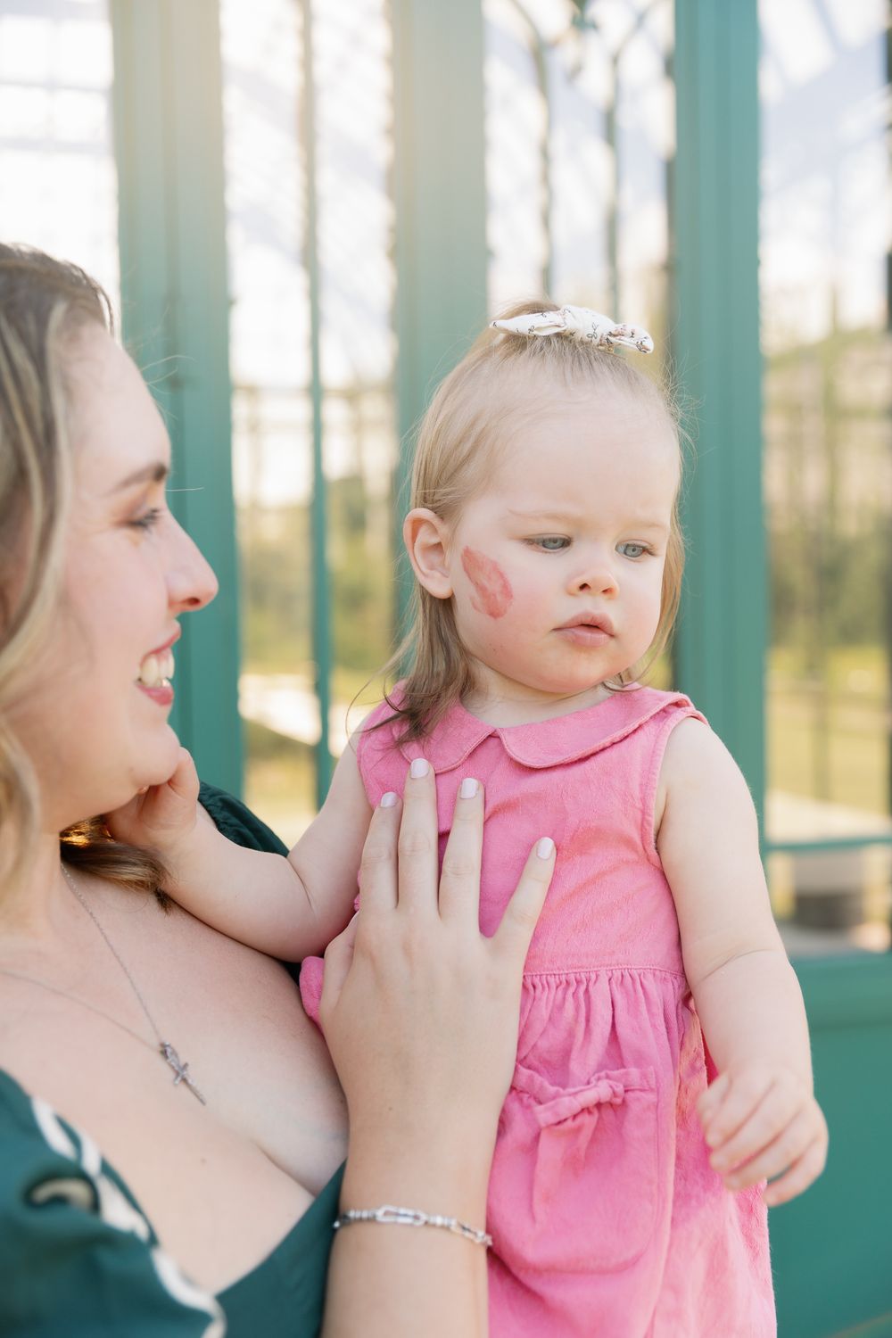 A mother holds her toddler girl in a pink dress with a lipstick kiss mark on her cheek outdoors.