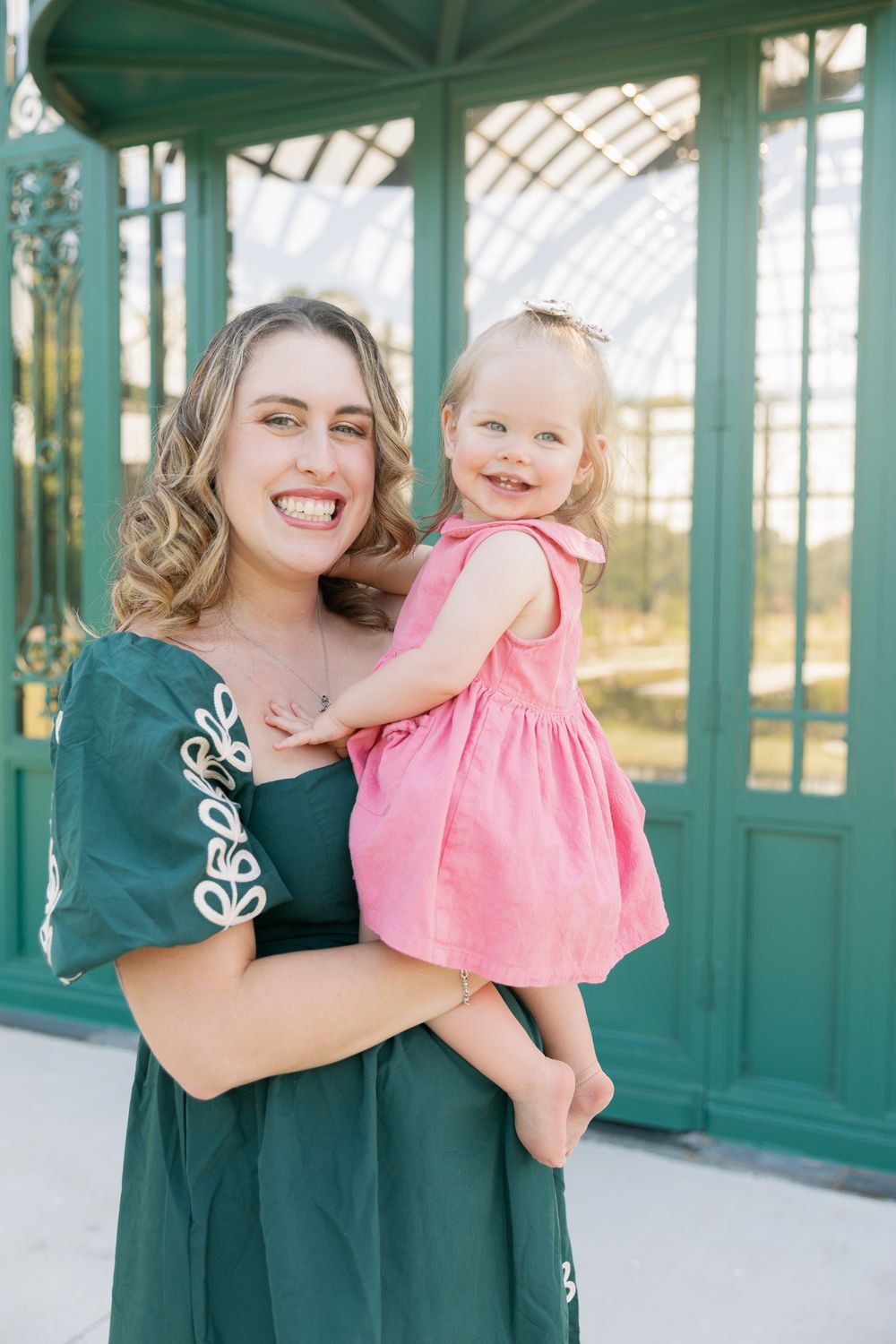 Smiling mother in green dress holds laughing toddler girl in pink dress outdoors near green glass building.