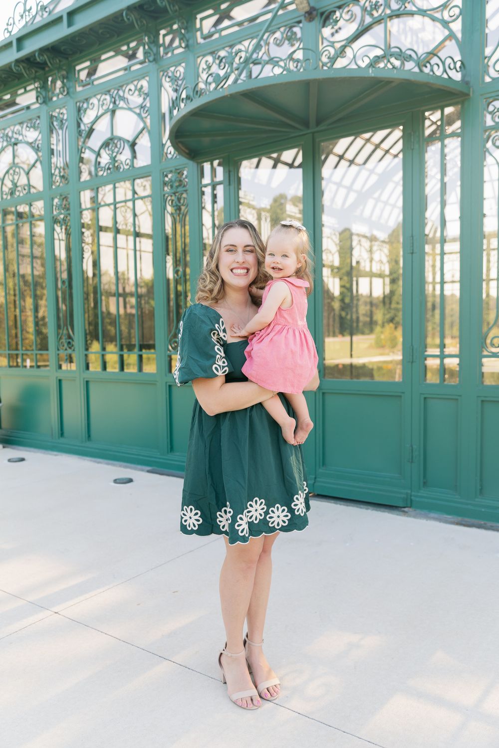Smiling woman in green floral dress holding toddler girl in pink dress outside ornate green greenhouse.