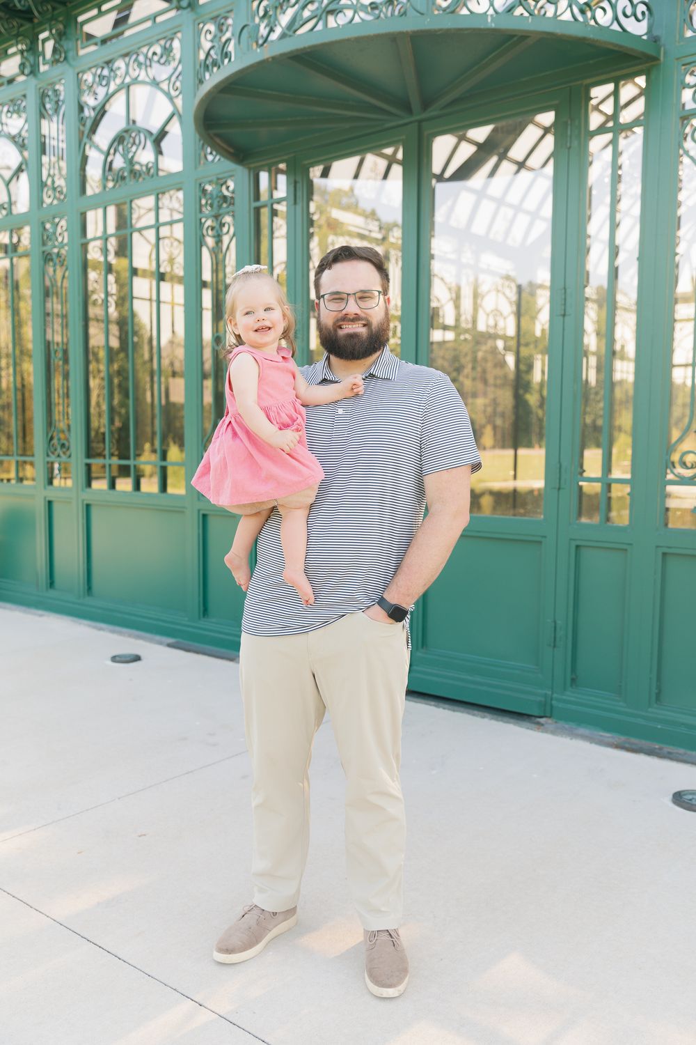 Man in striped polo holding smiling toddler girl in pink dress outside green ornate greenhouse.