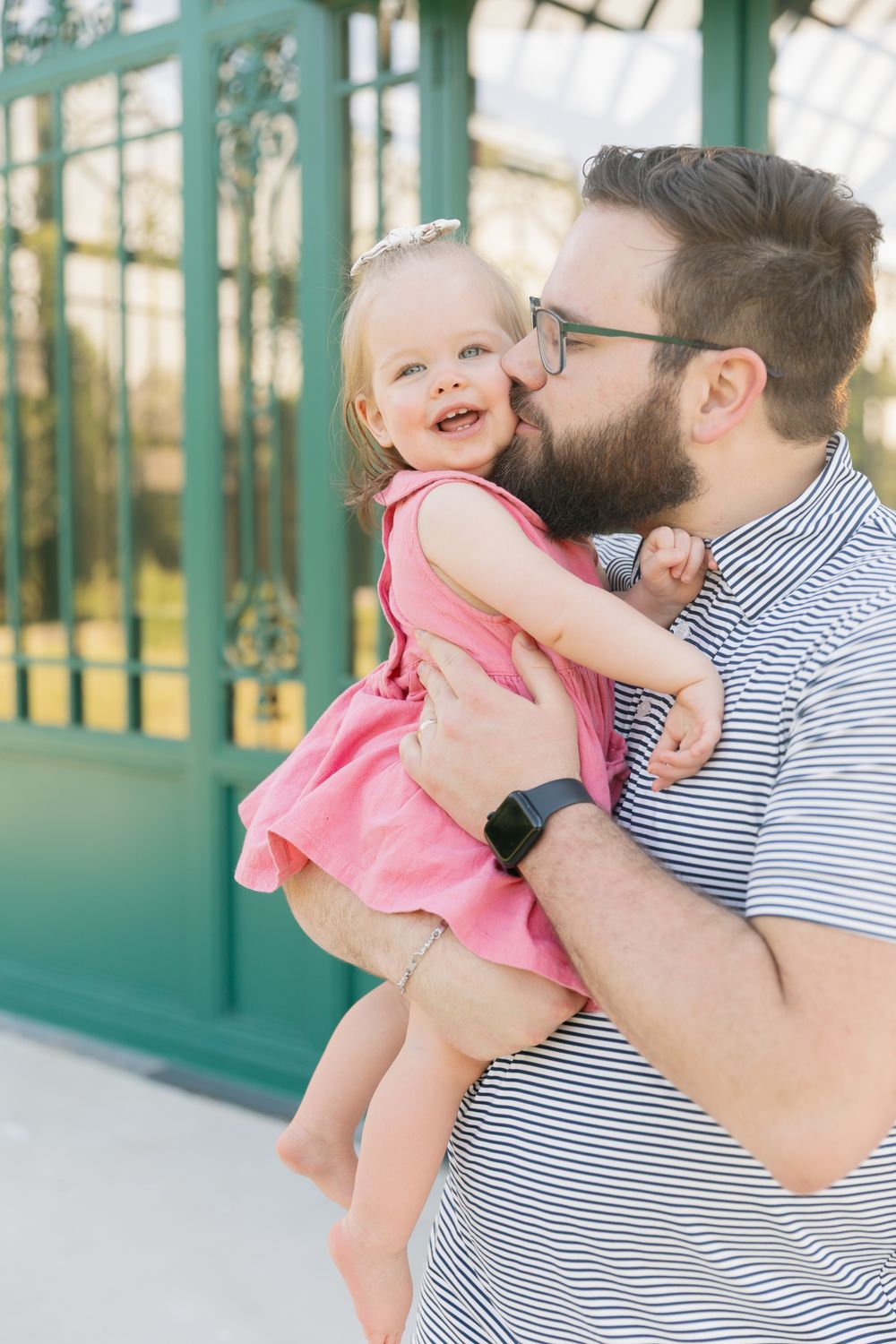Bearded father in striped shirt kissing smiling toddler girl in pink dress outdoors near green door.
