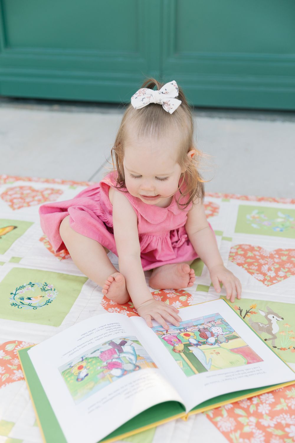 Toddler girl in pink dress with white bow reads a colorful picture book on a floral quilt outdoors.