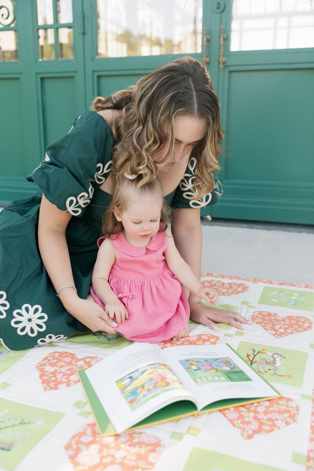 Mother in green dress reading colorful picture book with toddler girl in pink dress on floral quilt.
