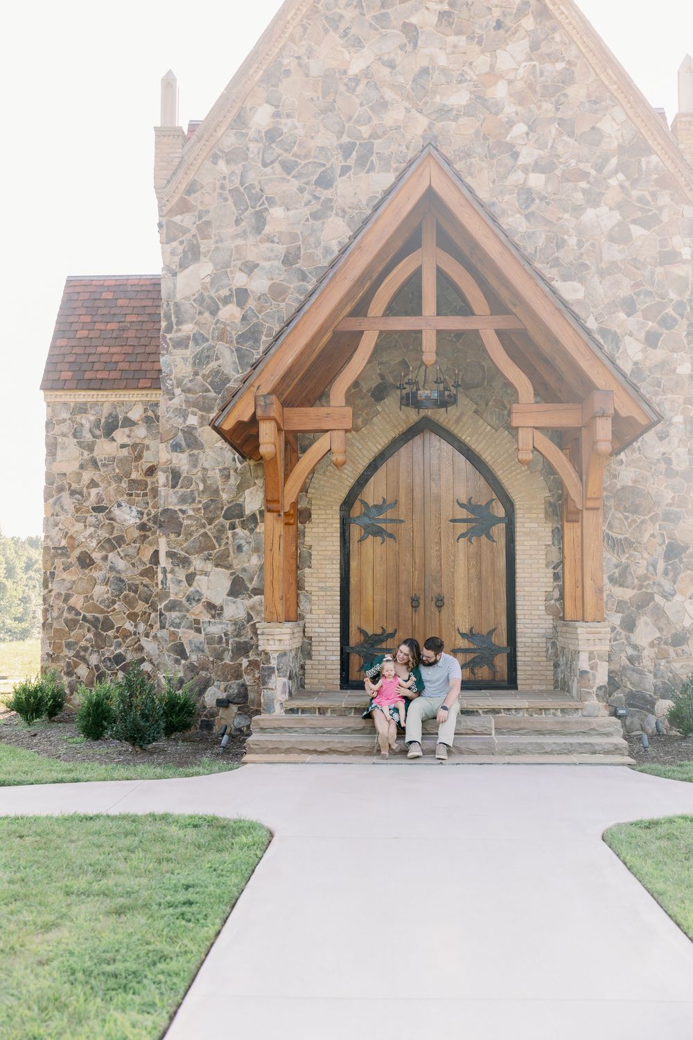 Family of three sits together on steps of rustic stone chapel with wooden arched door.