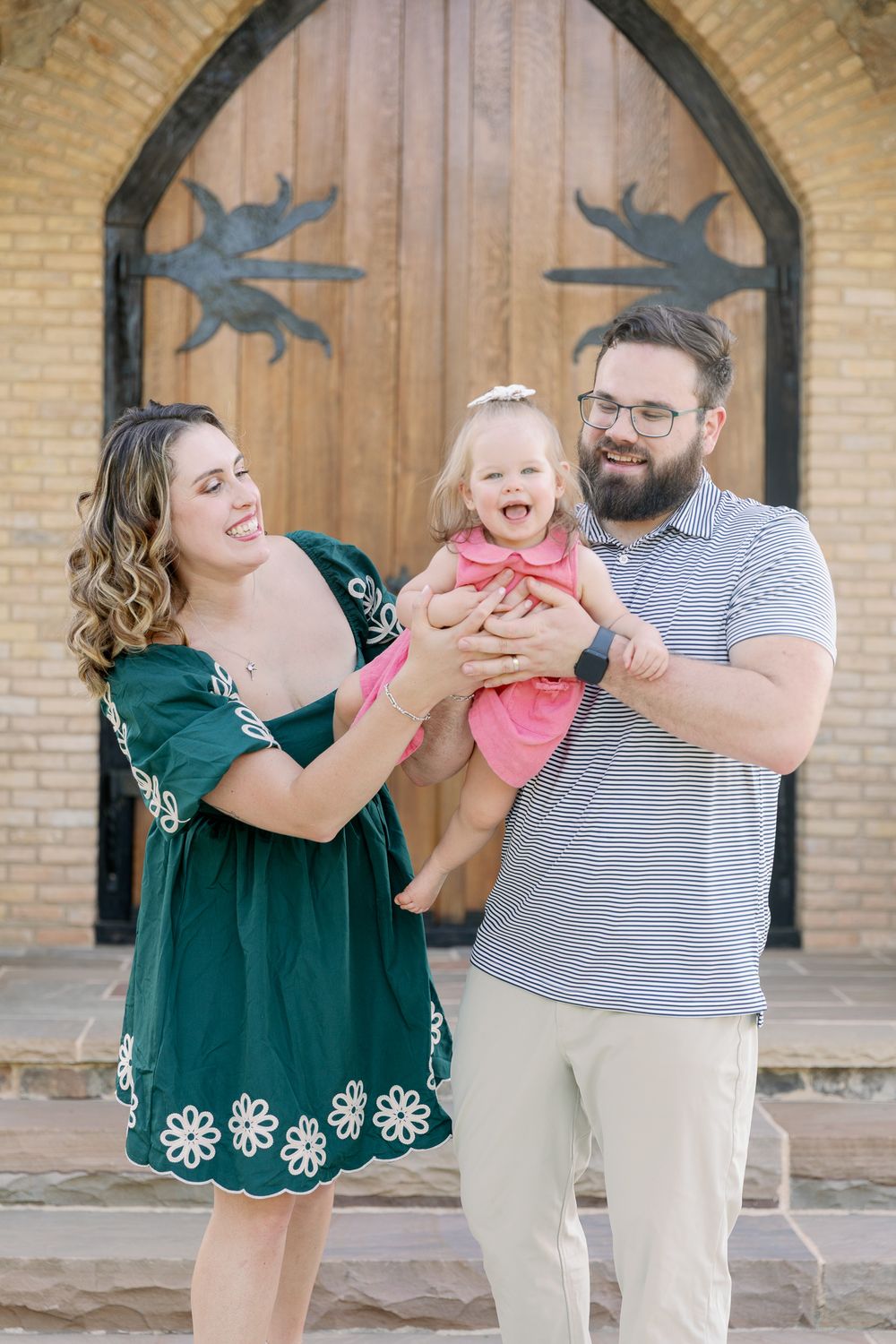 Happy family of three posing outdoors, parents holding laughing baby girl in pink dress before wooden church doors.