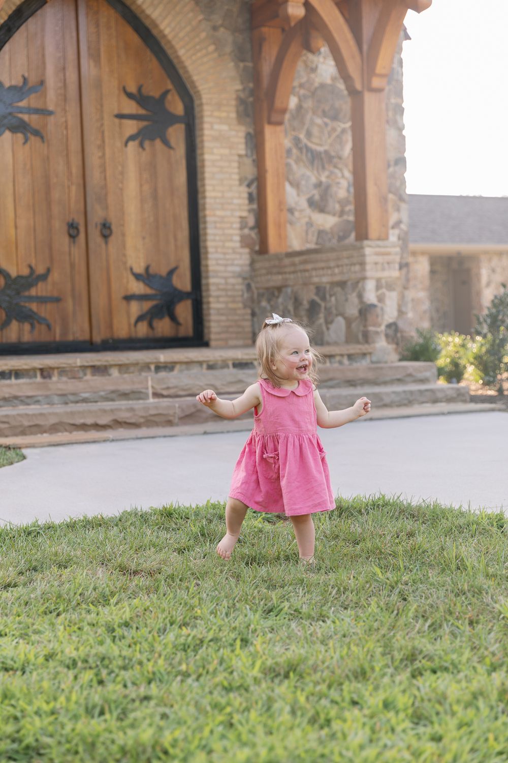 Happy toddler girl in pink dress laughing and walking on green grass outside rustic stone building.