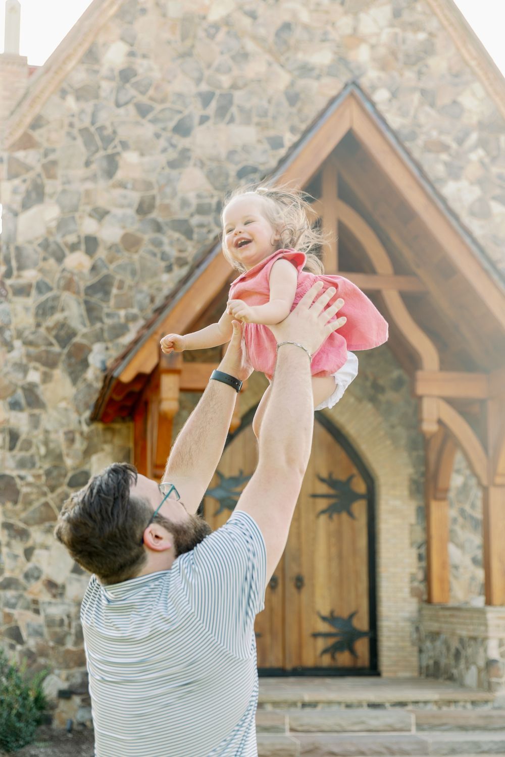Joyful father lifts laughing baby girl in pink dress high in the air outside stone church.