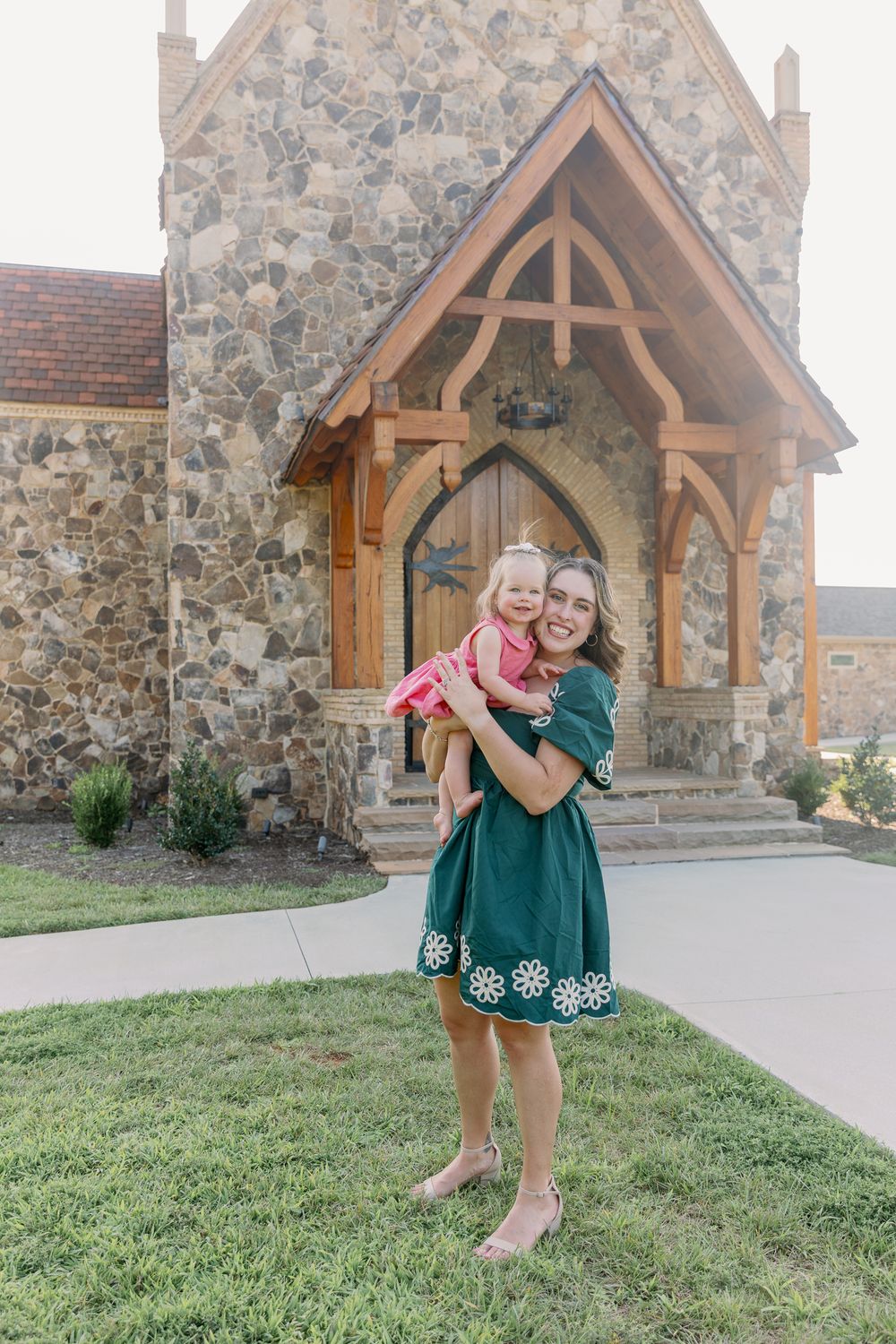 Woman in green dress holding a toddler girl in pink outside a stone chapel with wooden Gothic arch doorway.